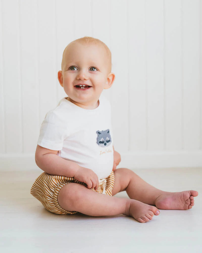 Baby sitting on a white surface wearing a Bebe white onesie with a bear design and gold bloomers.