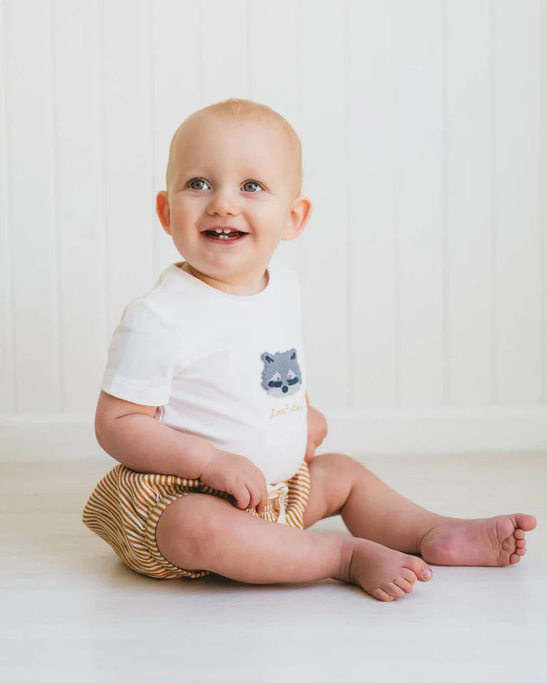 Baby sitting on a white surface wearing a Bebe white onesie with a bear design and beige shorts.