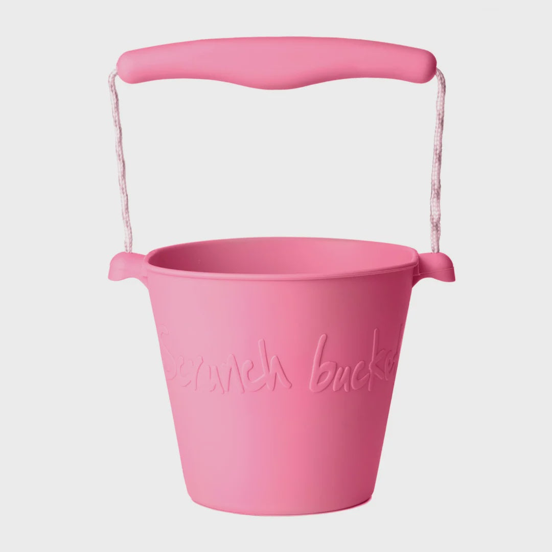 A pink plastic bucket with a matching pink handle and rope loop, featuring embossed script text on the front, shown against a light gray background.