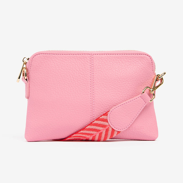 Pink crossbody bag with a red and white patterned strap on a light gray background