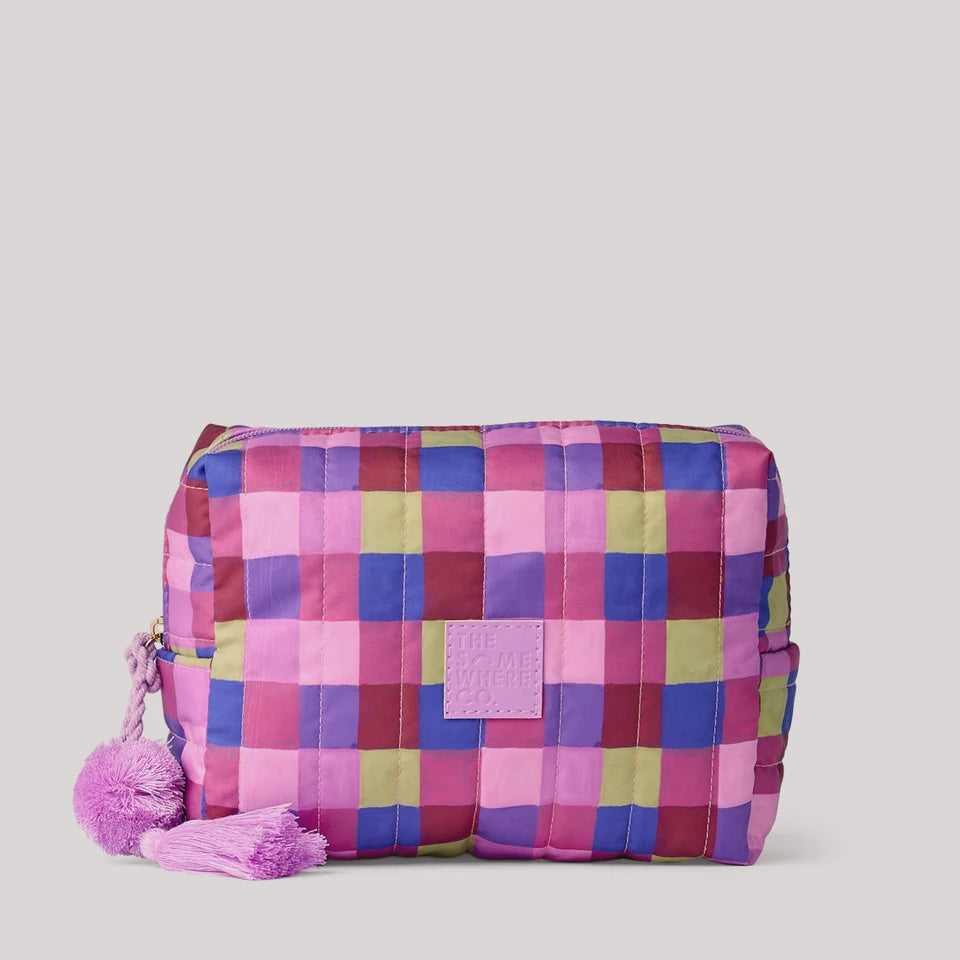 A quilted cosmetic pouch with a colorful plaid pattern in pink, purple, blue, and green, featuring a pink tassel zipper pull, set against a solid light gray background.