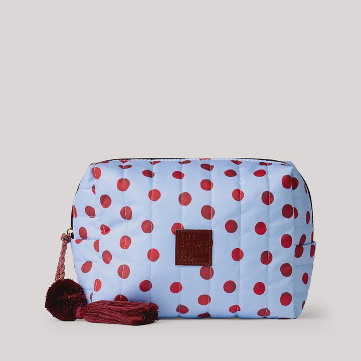 A light blue quilted cosmetic pouch with red polka dots, featuring a dark red leather patch on the front and a matching dark red tassel zipper pull, set against a solid light gray background.