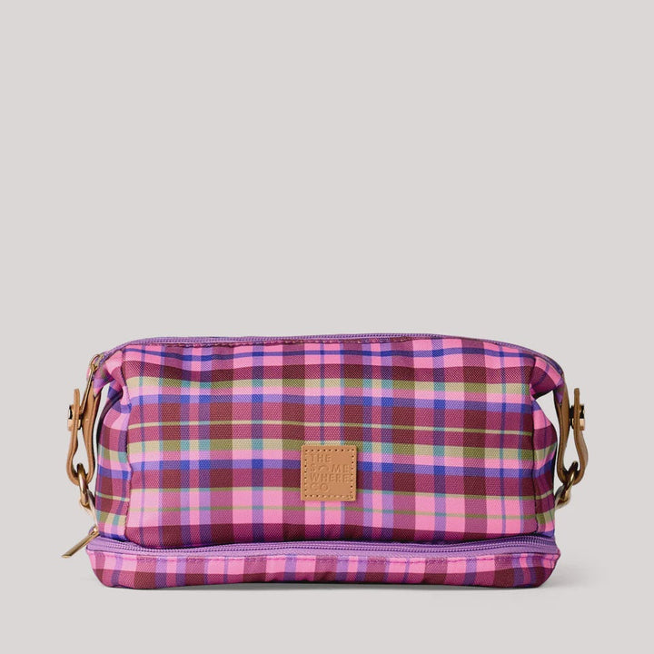 A small rectangular zip pouch with a pink, purple, and red plaid pattern, featuring a tan leather patch on the front and a gold-tone side strap, set against a light gray background.