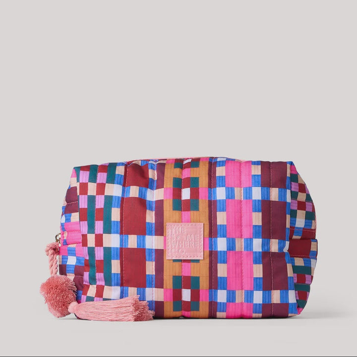 A cylindrical cosmetic bag with a colorful geometric patchwork pattern in pink, blue, red, and brown, featuring a pink tassel charm and a central pink brand patch.