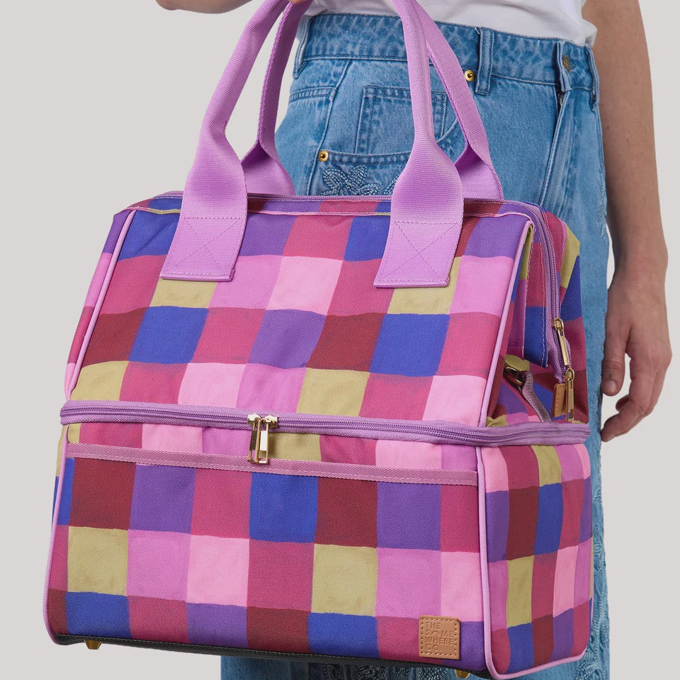 A person holding a colorful checkered zippered duffel bag with purple handles, shown against a light gray studio background.