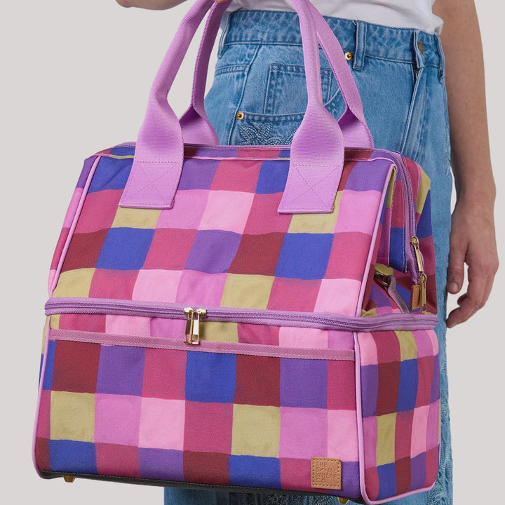 A person holding a colorful checkered zippered duffel bag with purple handles, shown against a light gray studio background.