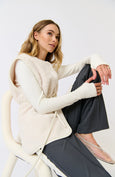 Woman wearing a cream cardigan over a dark outfit, sitting on a white chair against a plain background.