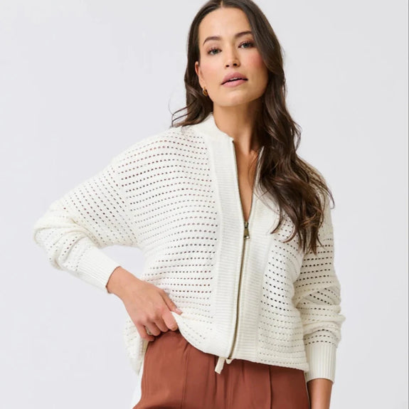 Woman wearing a white knit cardigan and brown pants on a light gray background