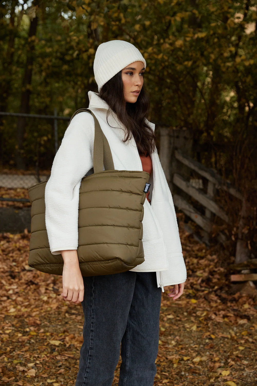 Person wearing a white jacket, green puffer vest, and white beanie outdoors with trees in the background.