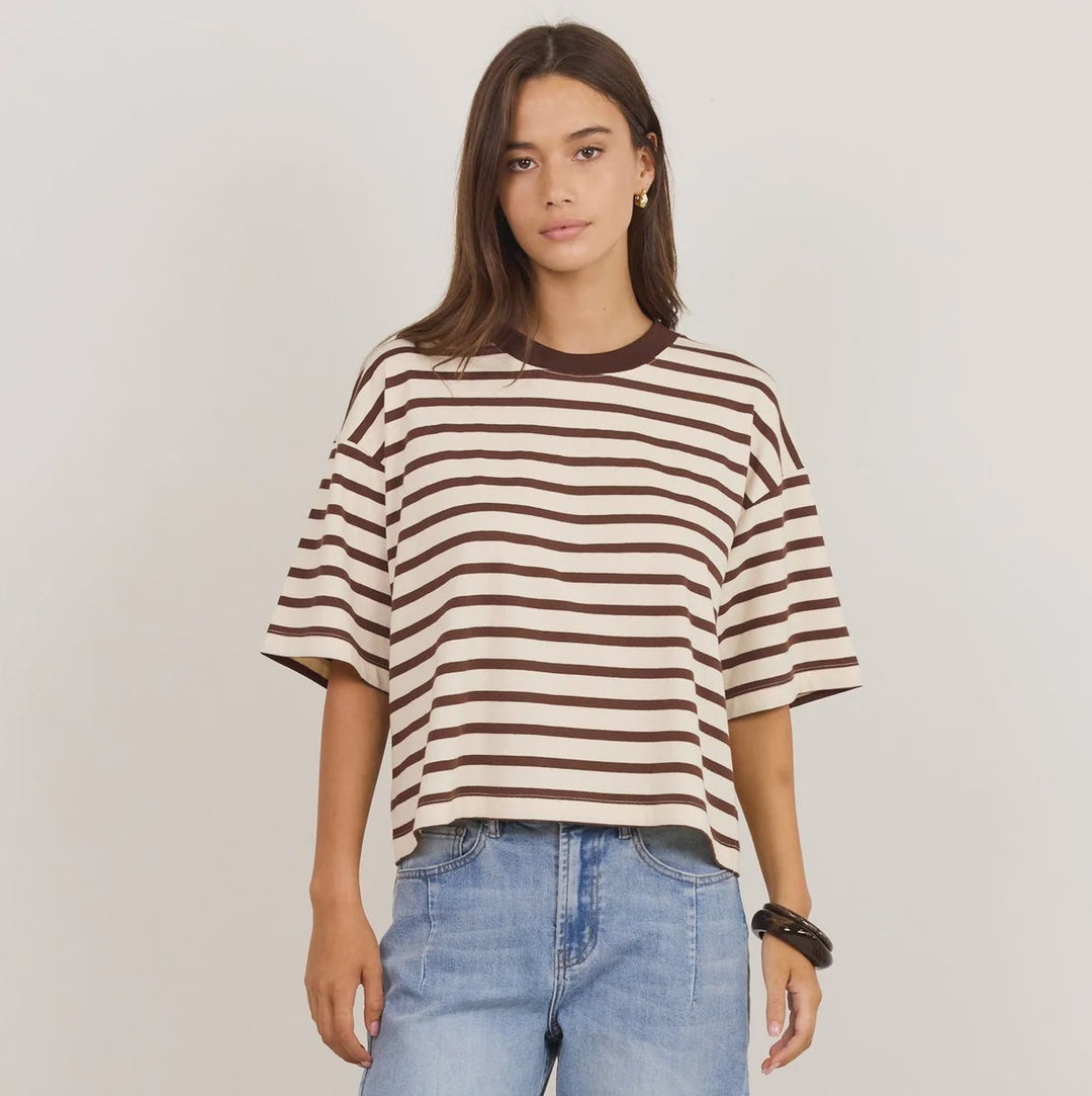 A woman modeling an oversized short-sleeve striped t-shirt in brown and cream, paired with light-wash baggy jeans and black sandals, against a plain light gray studio background.