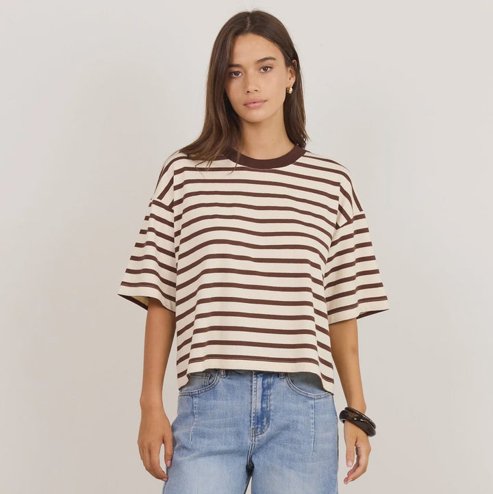 A woman modeling an oversized short-sleeve striped t-shirt in brown and cream, paired with light-wash baggy jeans and black sandals, against a plain light gray studio background.