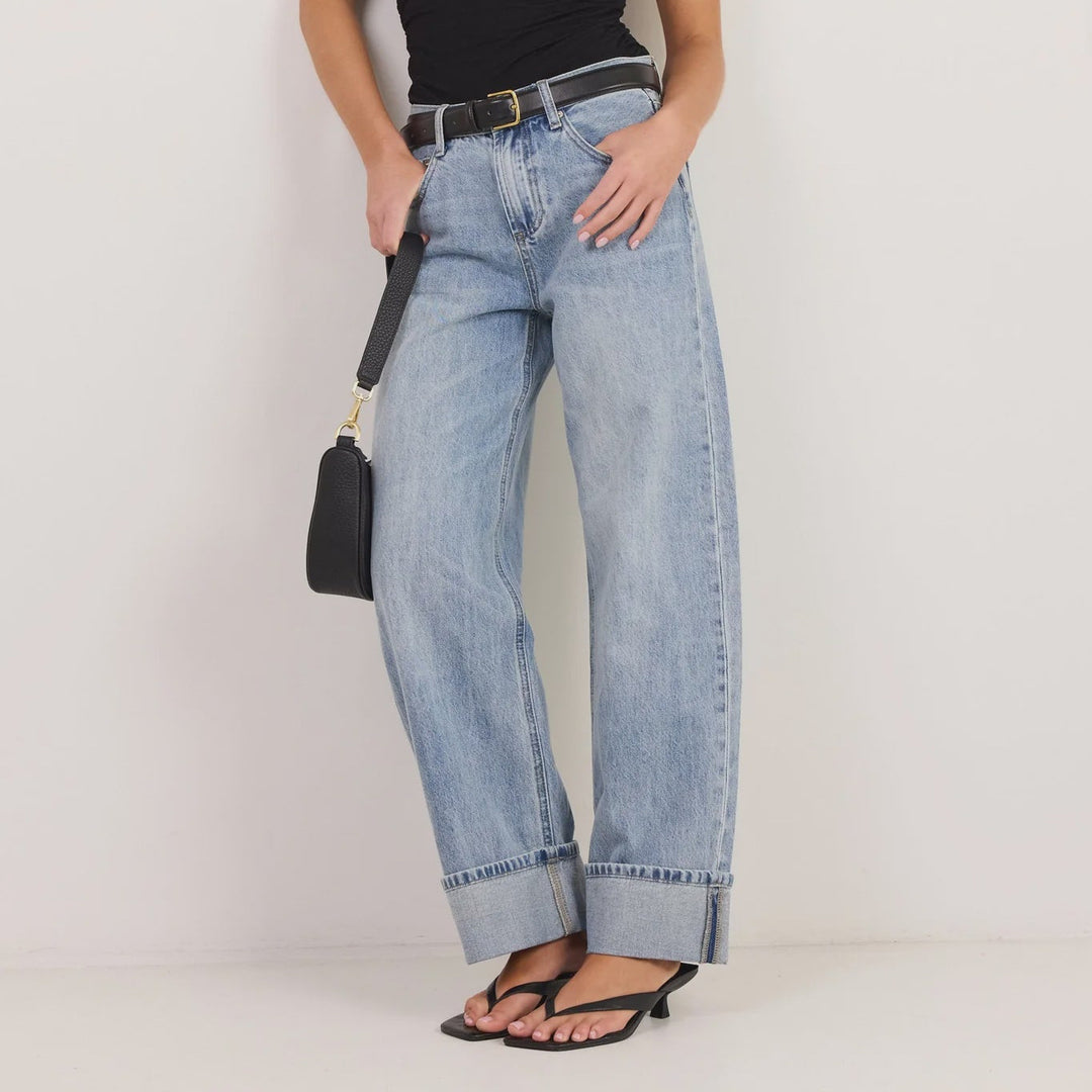 A woman modeling a black sleeveless wrap top, light-wash wide-leg denim jeans with rolled cuffs, black sandals, and a small black handbag against a plain white studio background.