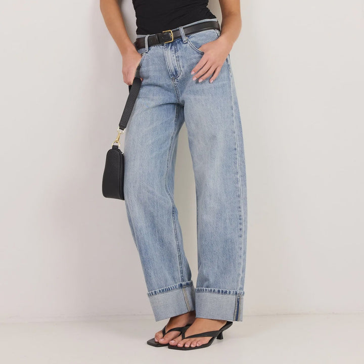 A woman modeling a black sleeveless wrap top, light-wash wide-leg denim jeans with rolled cuffs, black sandals, and a small black handbag against a plain white studio background.