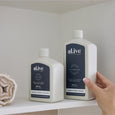 Two bottles of 'alive' hand and body products on a shelf with a hand holding one bottle.