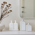 White ceramic bottles with 'aliver' branding on a shelf with a mirror and decorative plant.
