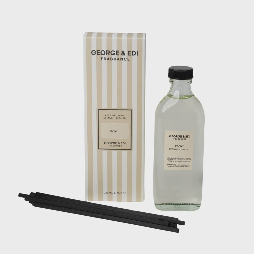 George & Edi Fragrance 'Envy' reed diffuser set, featuring a clear glass bottle with clear liquid, a striped cream and beige box, and four black reeds on a light gray background.