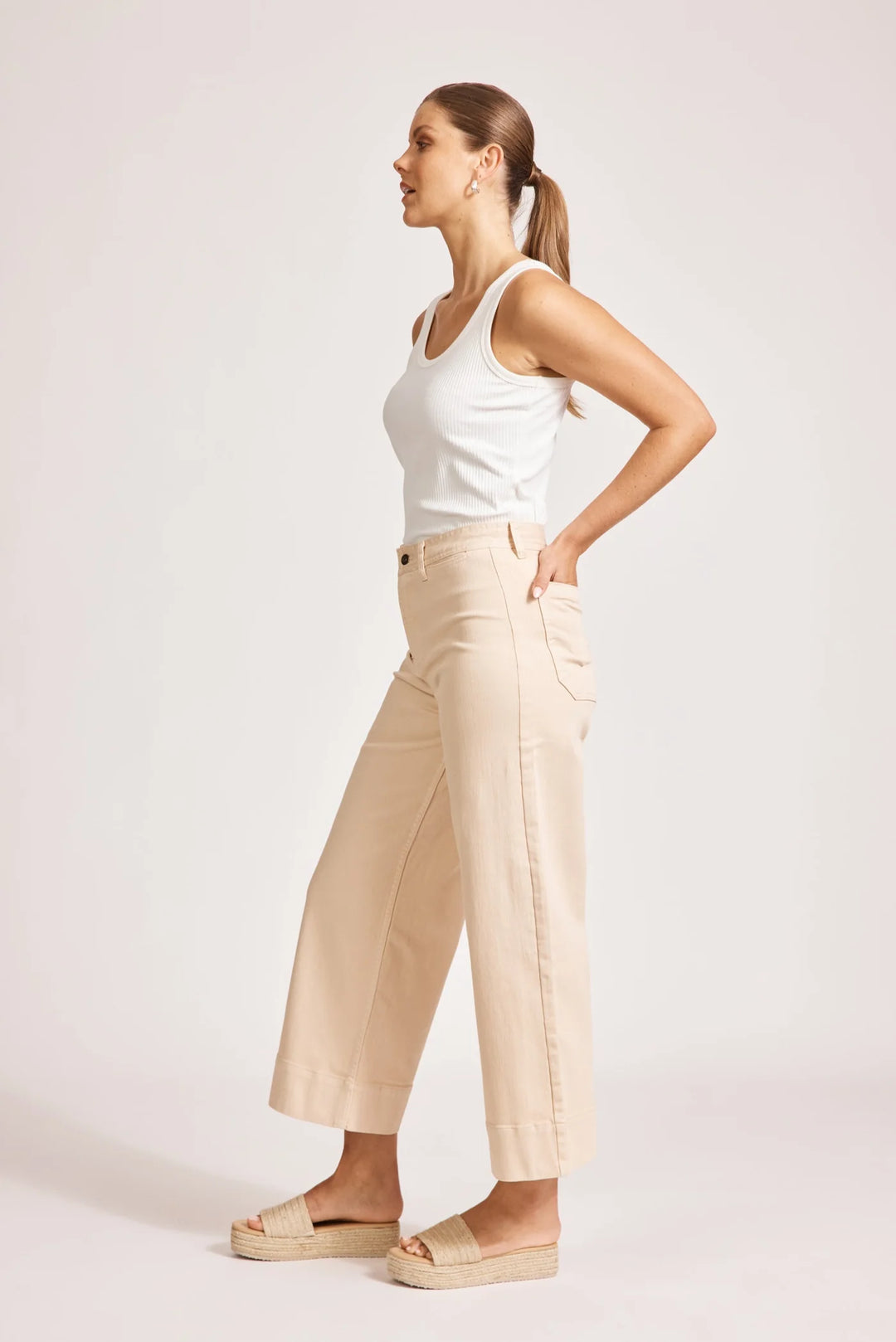 Woman wearing a white tank top and Eb & Ive beige pants on a light pink background