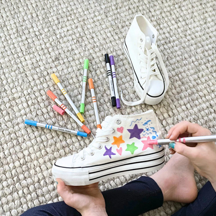 White sneakers with colorful star designs on a textured surface, surrounded by markers.