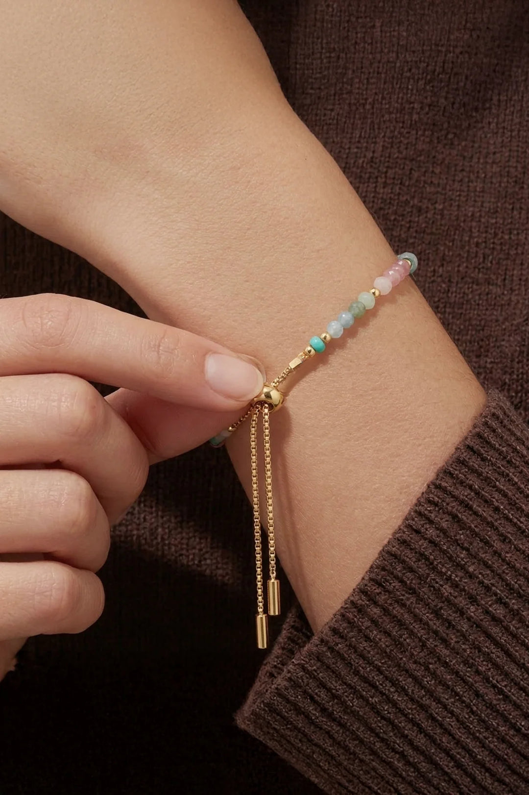 Close-up of a hand wearing a beaded bracelet with gold tassels on a brown background