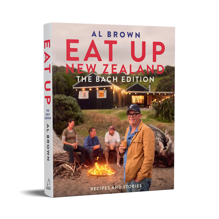 The book 'Eat Up New Zealand: The Bach Edition' by Al Brown, shown as a 3D paperback mockup against a clean white background. The cover features a photograph of a group of friends sitting around a campfire in front of a cabin at dusk, with the title in bold red and blue lettering.