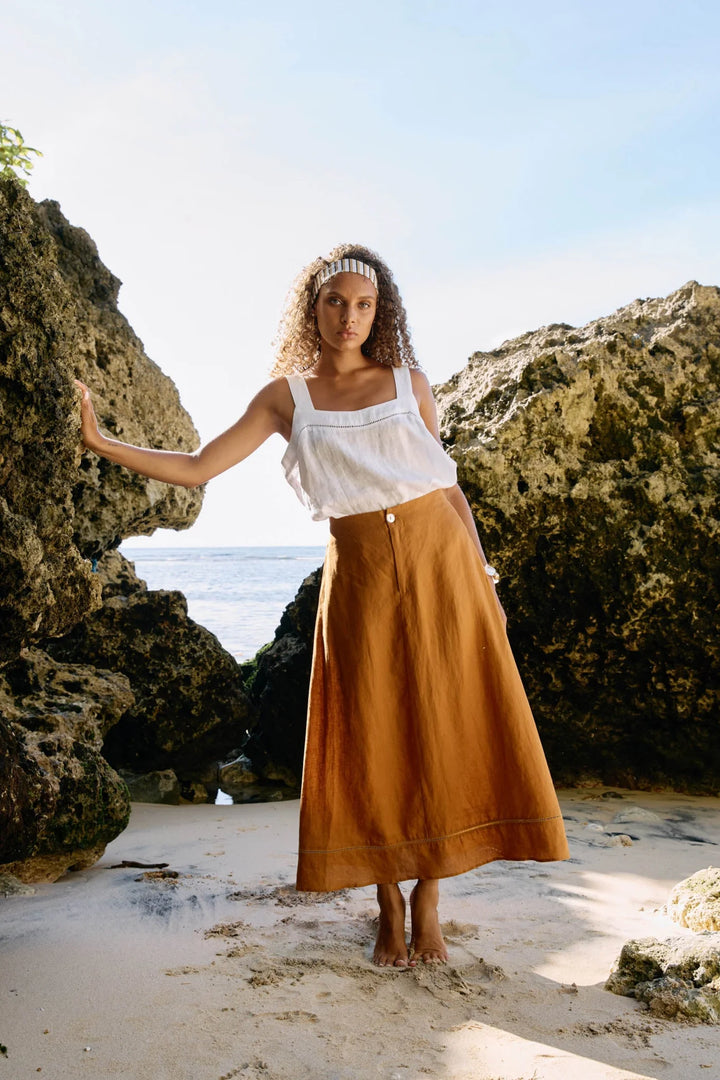 Woman in a white top and brown Eb & Ive skirt standing on a beach with rocky cliffs in the background.