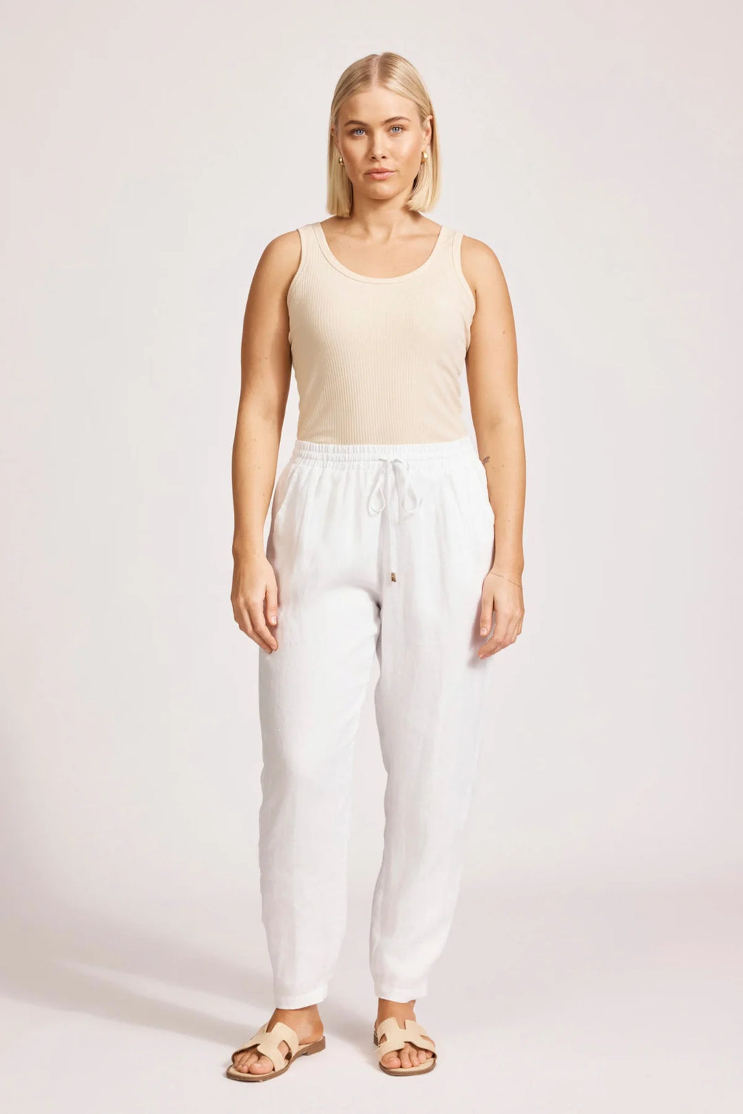 Woman wearing a beige tank top and eb & ive white pants on a plain background