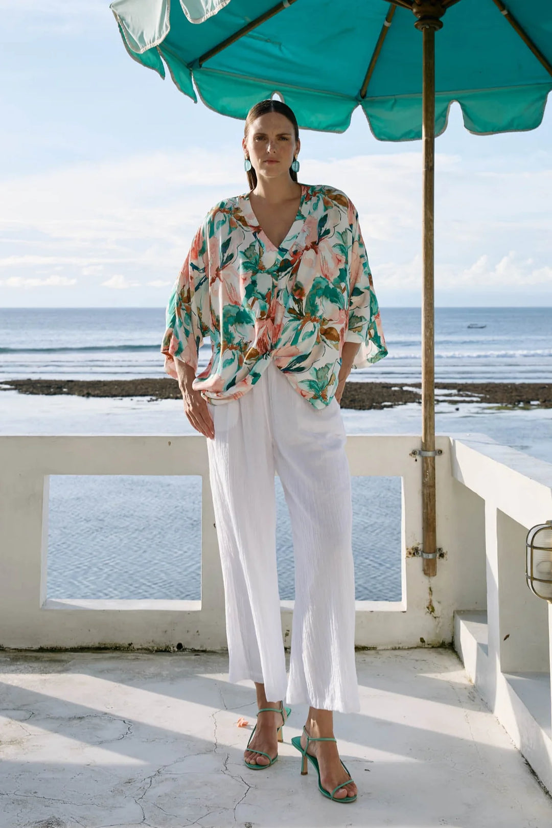 Woman in a floral blouse and Eb & Ive white pants standing under a teal umbrella by the ocean.