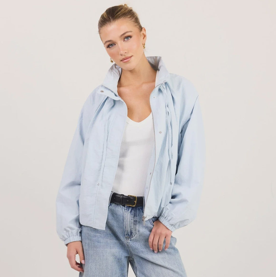 A woman modeling a light blue denim jacket and matching wide-leg jeans with a white tank top and black sandals, standing against a plain off-white studio background.