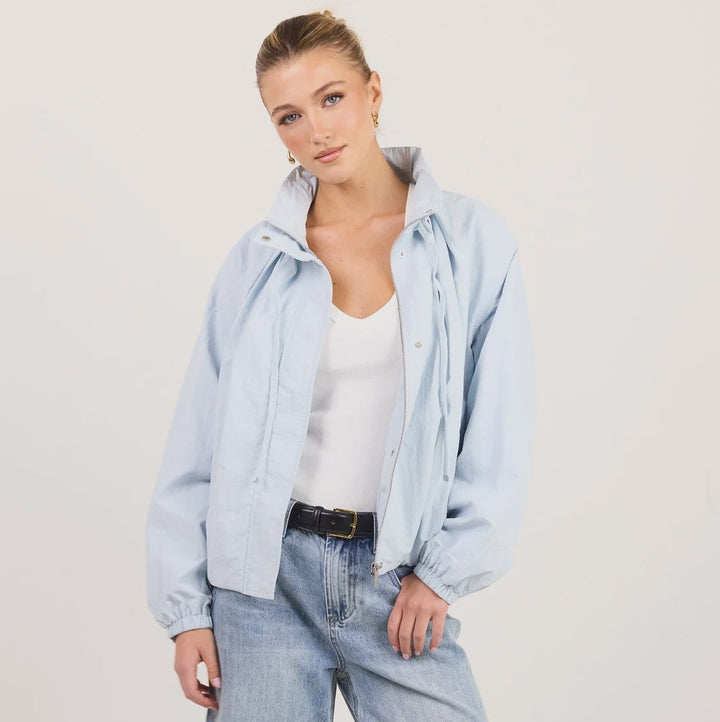 A woman modeling a light blue denim jacket and matching wide-leg jeans with a white tank top and black sandals, standing against a plain off-white studio background.