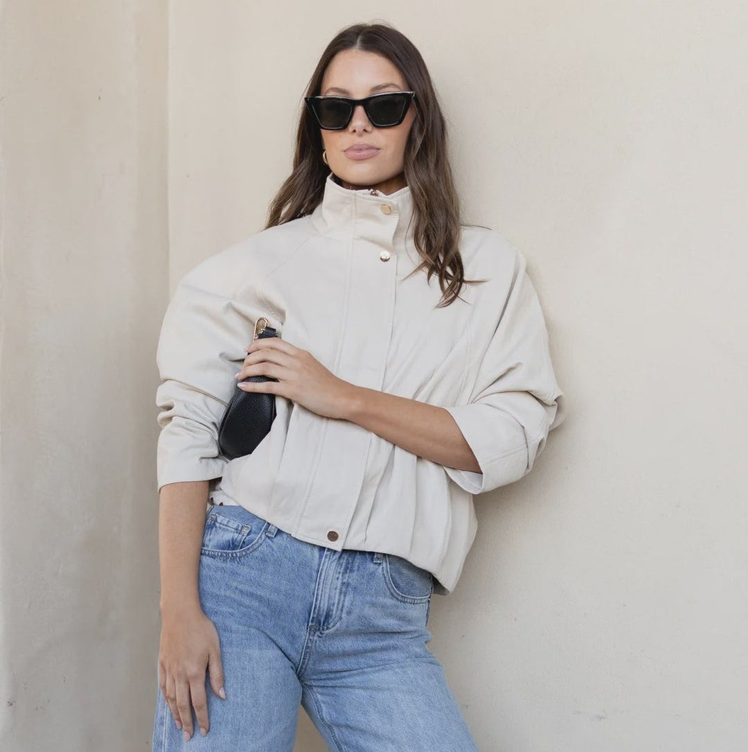 A woman modeling a white long-sleeve turtleneck top and light blue wide-leg cropped jeans, standing against a beige stucco wall on a brick floor.