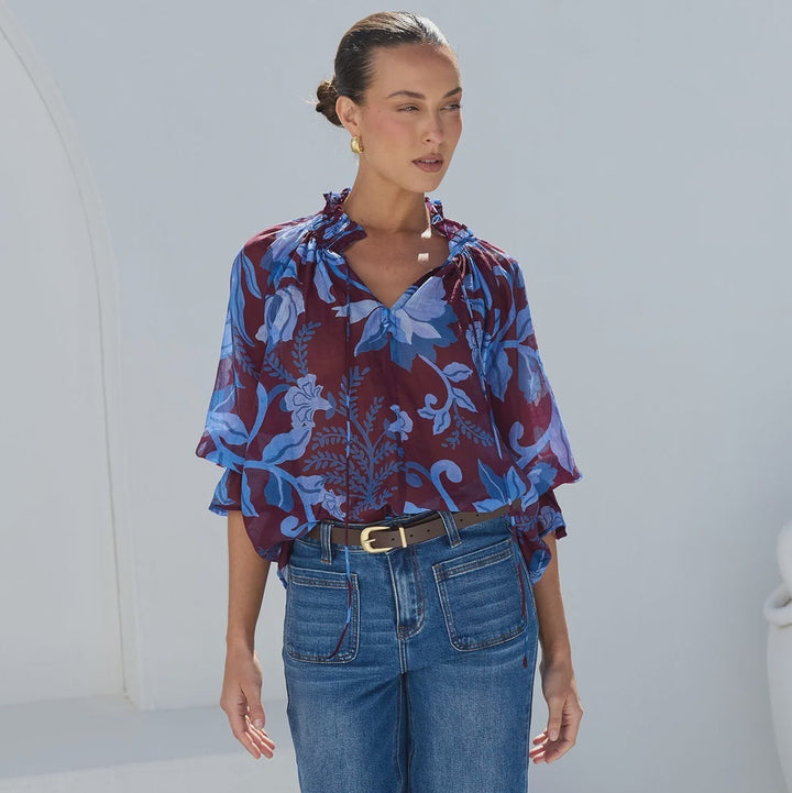 A woman modeling a blue and purple abstract patterned blouse with ruffled sleeves, tucked into wide-leg blue jeans, standing in front of a white wall.
