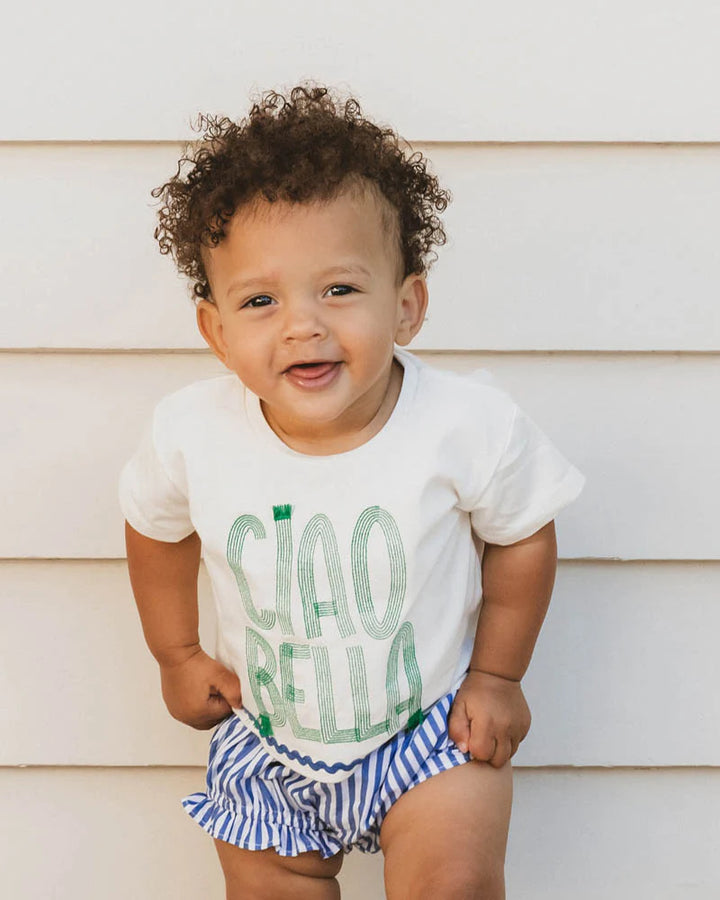 Baby wearing a white shirt with green text and striped diaper cover, standing against a light wooden wall.