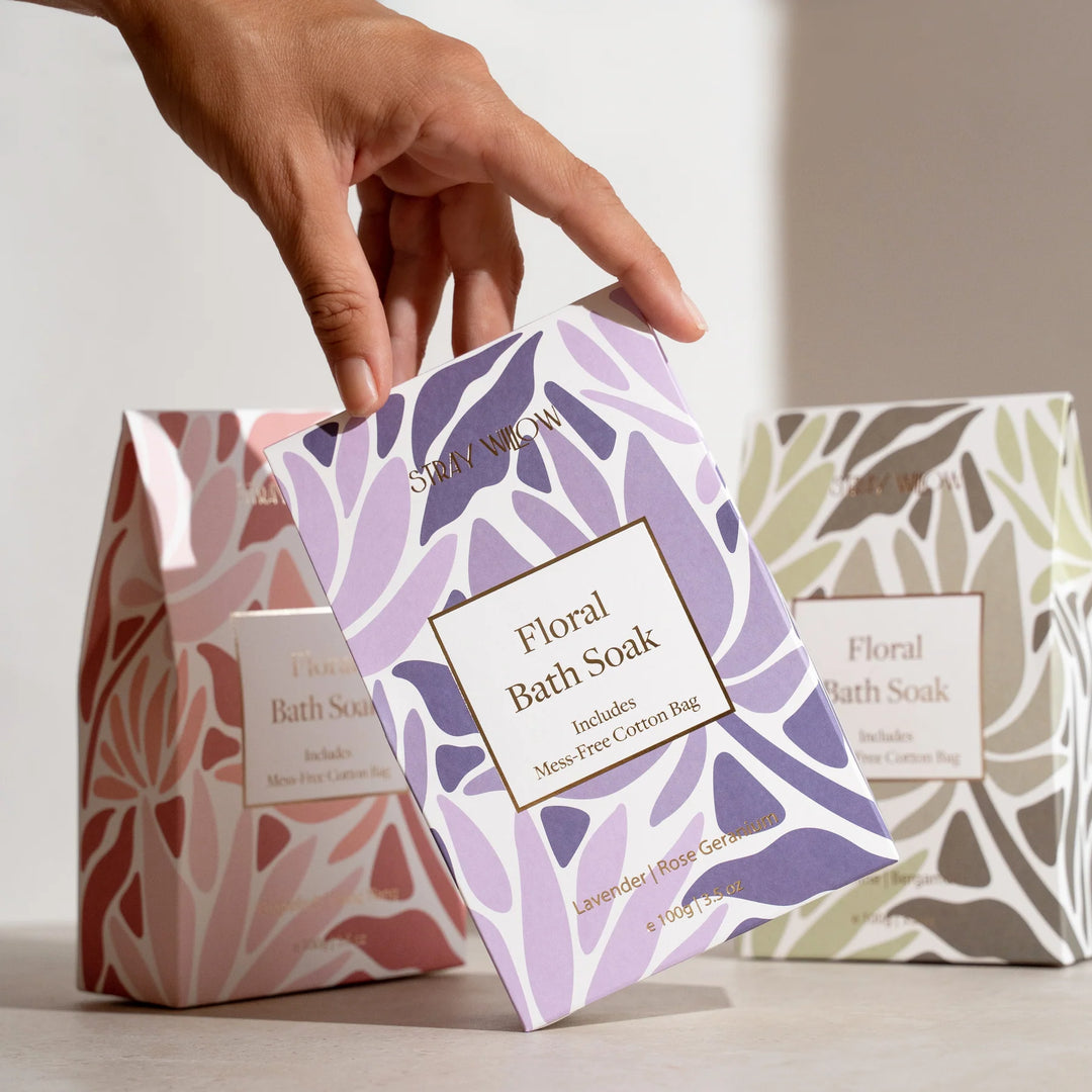 Hand holding a floral bath soak packaging with another one in the background on a light surface.