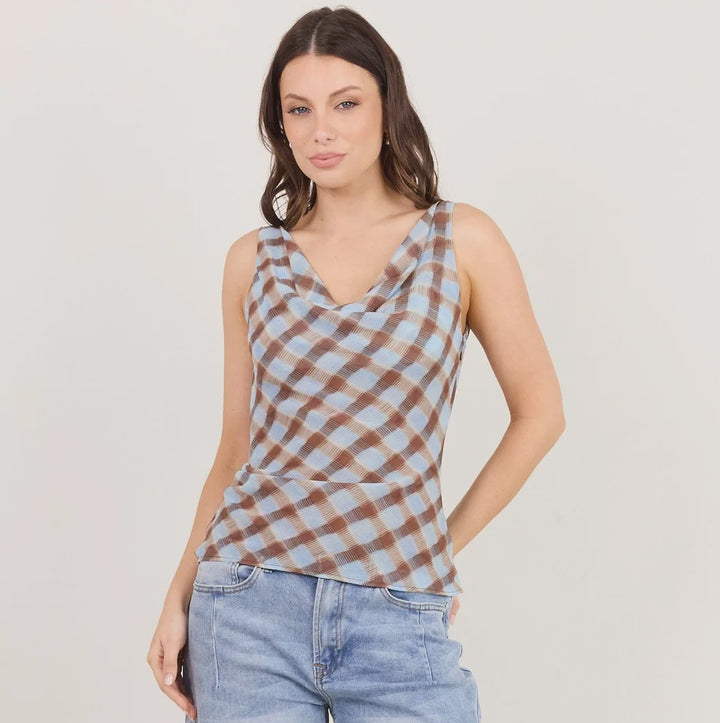 A woman modeling a sleeveless cowl-neck top with a brown and beige checkered pattern, paired with light-wash wide-leg jeans and black heeled sandals, against a plain off-white studio background.