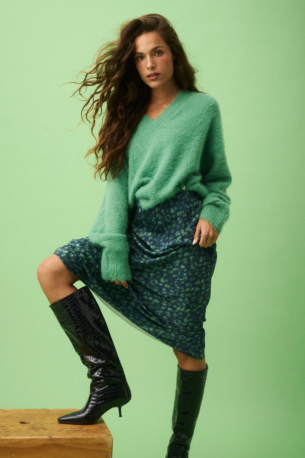 Woman wearing a green sweater and patterned skirt with black knee-high boots on a light green background