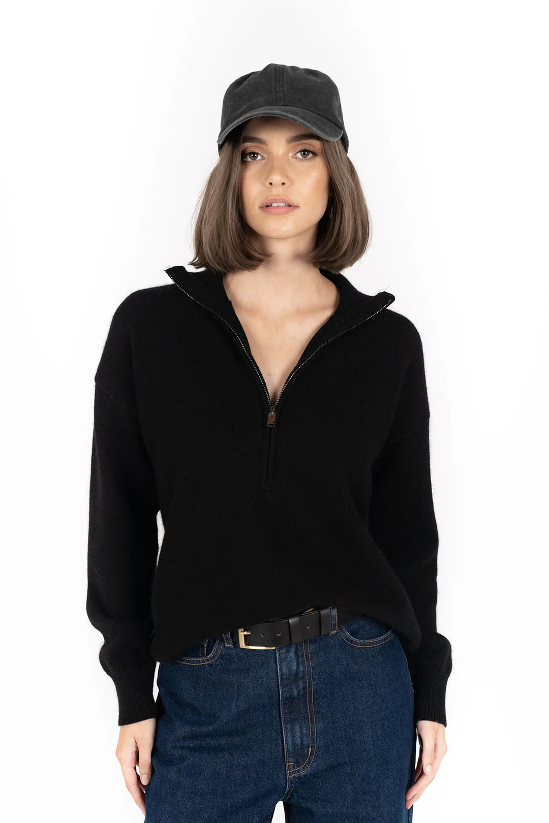 Person wearing a black zip-up hoodie, dark jeans, and a black cap on a white background