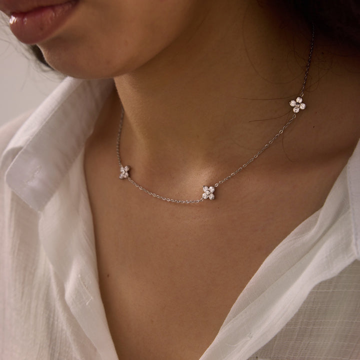 A close-up of a delicate silver chain necklace with three small white flower charms, worn by a model in a white collared shirt.
