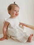 Baby in a white dress sitting on a white surface with a wooden toy.