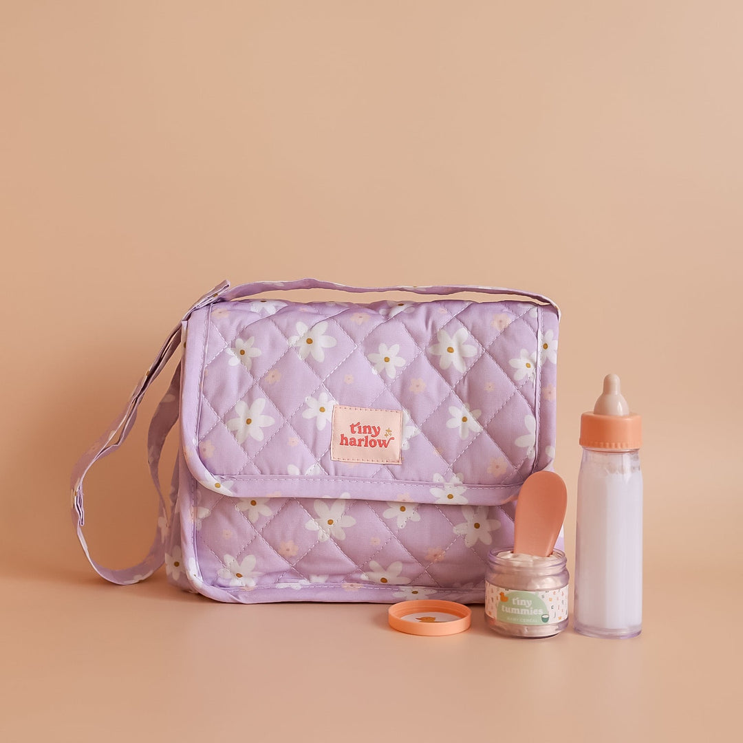 A lavender quilted diaper bag with a white daisy floral pattern, shown with baby accessories including a baby bottle, a small jar of powder with a scoop, and a small orange ring on a solid peach-beige background. The brand label on the bag reads 'tiny harlow'.