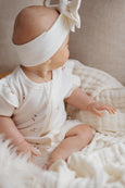 Baby in a white outfit with a bow headband sitting on a soft surface.