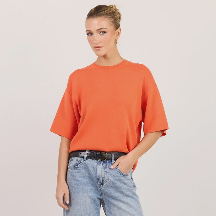 A woman modeling a bright orange short-sleeve crewneck top and light-wash wide-leg blue jeans with rolled cuffs, paired with a black belt and black sandals, against a plain off-white studio background.