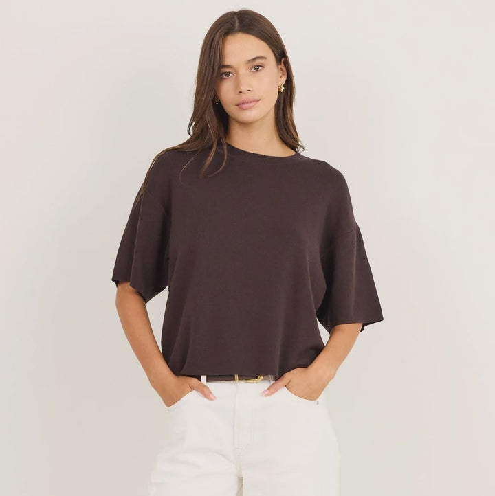 A woman modeling a dark brown short-sleeve top and white wide-leg pants against a plain white studio background.