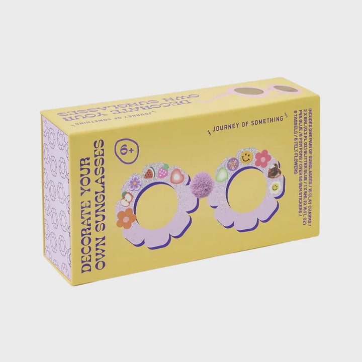 A yellow cardboard box for 'Journey of Something' eyeglass decoration kits, featuring illustrations of colorful flower and pom-pom decorated glasses on a white background.