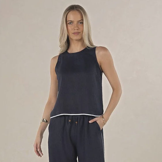 A woman modeling a sleeveless navy blue jumpsuit with white contrast piping, standing against a neutral beige studio background.
