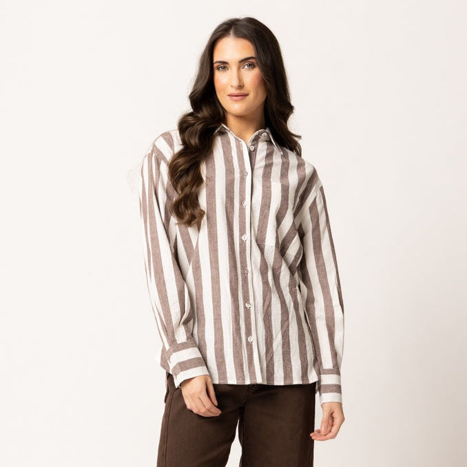 A woman modeling a brown and beige vertically striped button-down shirt paired with dark brown wide-leg trousers and black pointed-toe flats, set against a plain off-white studio background.