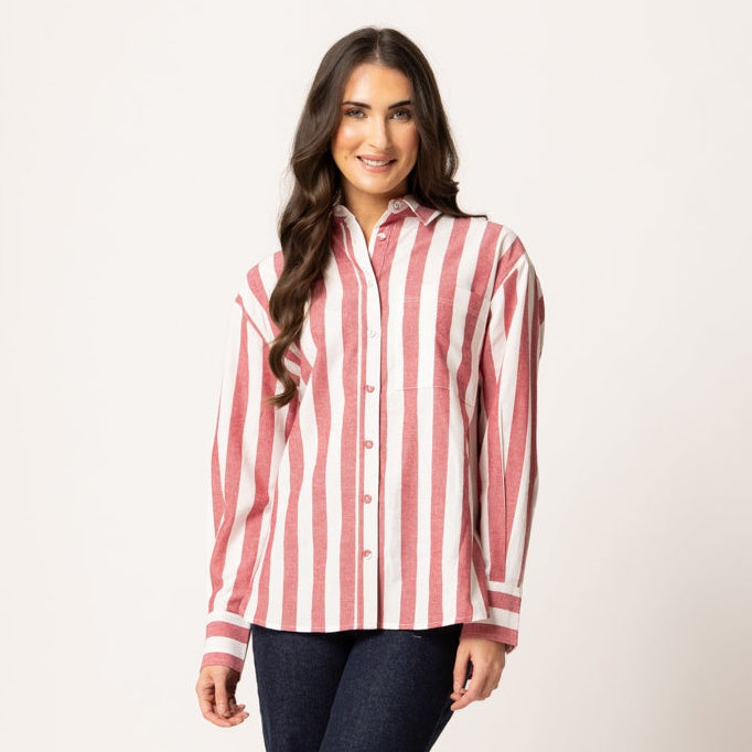 A woman modeling a long-sleeve button-up shirt with vertical pink and white stripes, paired with dark blue flared jeans and beige sandals, against a plain light background.