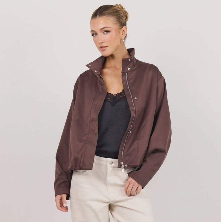 A woman models a dark brown satin bomber jacket over a black top and cream-colored wide-leg trousers, standing against a plain white studio background.