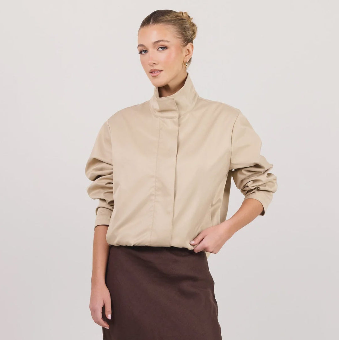 A woman modeling a beige mock-neck long-sleeve blouse and a dark brown midi skirt, standing against a plain white studio background.