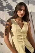 Woman wearing a light yellow sleeveless top against a textured wall.