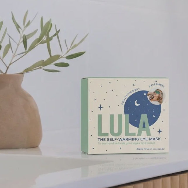 A box of LULA The Self-Warming Eye Mask sits on a white countertop next to a beige ceramic vase with green leaves, in a bright, modern bathroom setting.